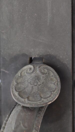 Close-up of an antique bronze entry door pull with a decorative round thumb latch plate and visible leafy handle detail.