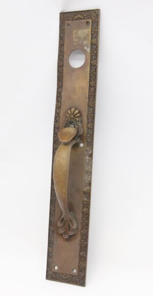 Antique bronze entry door pull with a flower and shell design, shown as a tall rectangular plate with a thumb latch, lock opening, and visible bend.