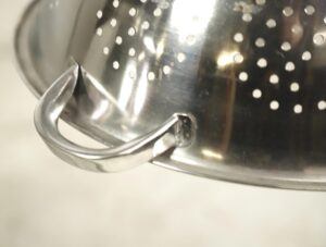 Close-up of a stainless steel colander pendant light with a curved handle and perforated surface.