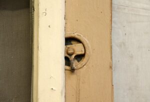 Close-up of an old screen door latch beside the screen, showing chipped tan paint and worn hardware.