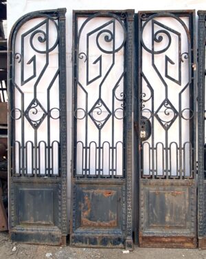 Art Nouveau wrought iron bifold doors with decorative scrollwork and plain lower panels, shown closed with visible cast iron trim and worn areas at the bottom.