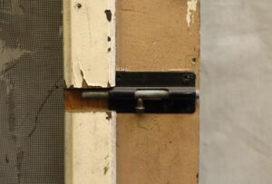 Close-up of an old screen door hinge and latch area with worn tan-painted wood, intact hardware, and the screen visible at the edge.