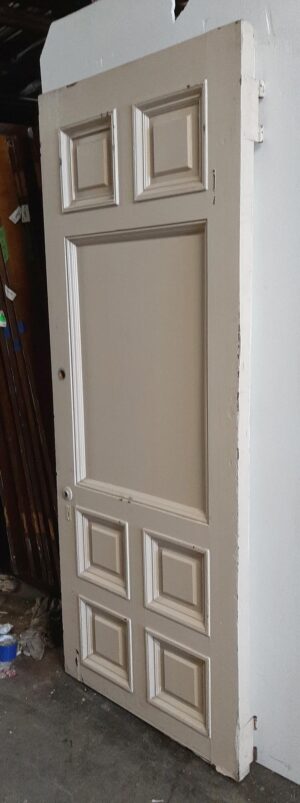 Tan wood entry door with seven raised panels and visible edge wear, shown at an angle.