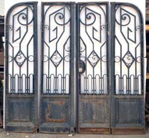 Front view of an Art Nouveau wrought iron bifold door set with four decorative panels, arched scrollwork, and plain lower panels showing wear and rust.