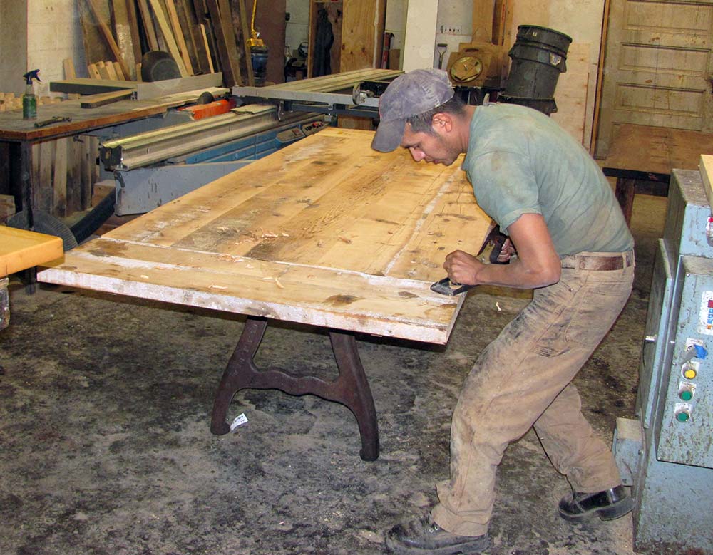 Hand planing A man in a cap sands a large tabletop in a cluttered workshop, with tools and lumber around him—demonstrating skilled craftsmanship as he repurposes old wood into furniture.