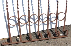 Salvaged wrought iron railing section with twisted vertical bars, circular loops, and small floral details, shown in a rusted finish.