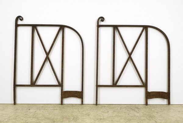 Pair of wrought iron hand railings shown side by side, each with an arched top rail, X-brace panels, and a curled end detail, viewed upright against a plain white backdrop.