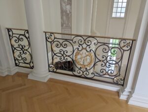 Decorative stair bannister panel with black wrought steel scrollwork, gilded brass accents, and a brass handrail, shown as a close-up section with a monogrammed center detail.