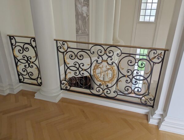 Decorative stair bannister panel with black wrought steel scrollwork, gilded brass accents, and a brass handrail, shown as a close-up section with a monogrammed center detail.