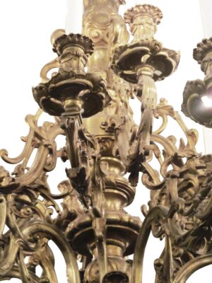 Close-up of an ornate 18-arm chandelier with scrolling rococo detailing, ornate bobeches, and candle-style sockets with white sleeves.
