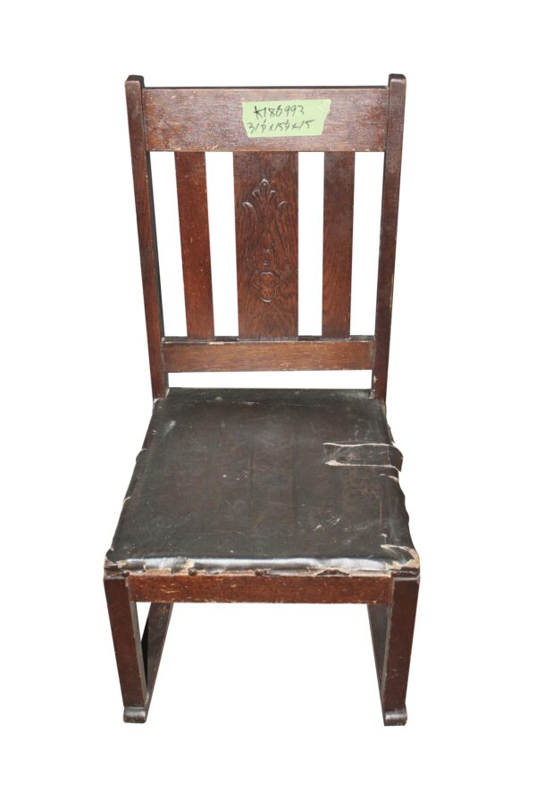 Arts and Crafts wooden chair with a dark wood frame, worn black seat, and carved back panel shown from the front.
