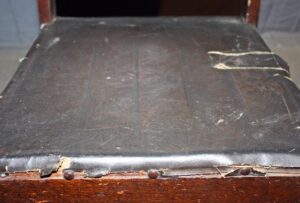 Close-up of the worn seat on an Arts & Crafts wooden rocker, showing dark wood and black upholstery with visible age and damage.