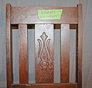Close-up of a dark wood Arts and Crafts rocker back with vertical slats and a carved center panel, shown with a green tape label near the top rail.