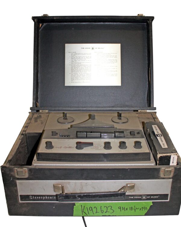 Open black metal portable reel-to-reel recorder with the lid raised, showing the control panel, reels, and instruction sheet inside.