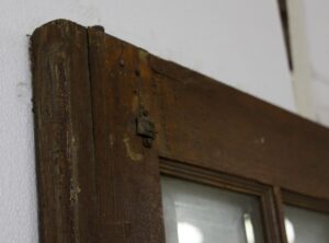 Close-up of the top corner of a dark wood tone French door with a beveled glass lite and worn antique hardware.