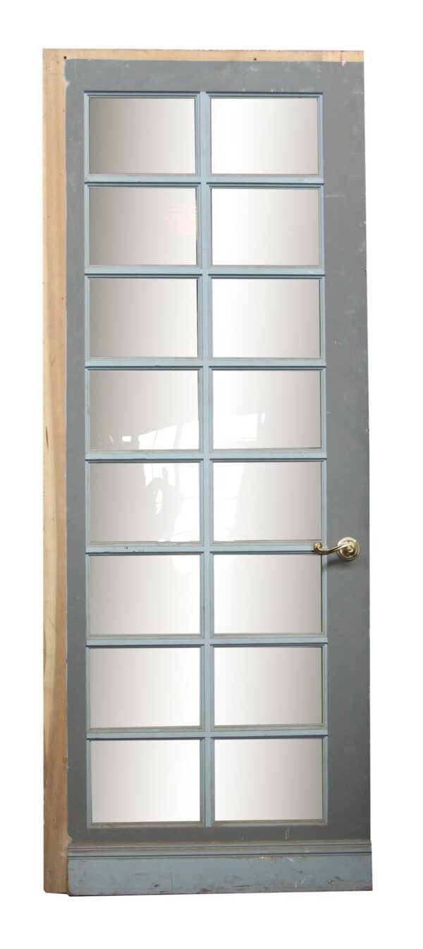 Vintage French door with 16 rectangular panes, painted gray on one side and fitted with a brass handle.