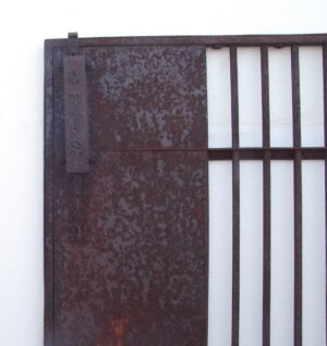 Close-up of an antique iron elevator gate with a solid lower panel, vertical bars on the right, and the original brass knob at the left edge, showing visible surface deterioration on the panel.