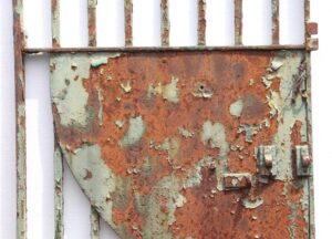 Distressed wrought iron gate with split finials and a rusted security panel, shown in close-up with peeling green paint.