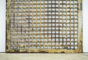 Reclaimed cast iron wall grate with a square lattice, surface rust, and chipped white paint.