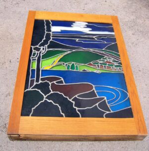 Stained glass window panel in a dark wood light box, showing a pastoral landscape with hills, sky, and water in multiple colors.
