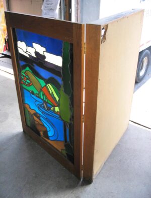 Backlit stained glass window mounted in a dark wood light box, shown at an angle with a multicolor pastoral scene and the side panel visible.