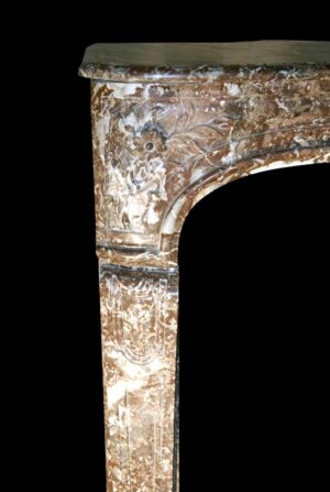 Close-up of a Louis XV marble mantel showing the left corner, with carved floral detail, an arched opening, and a mottled brown and white surface.