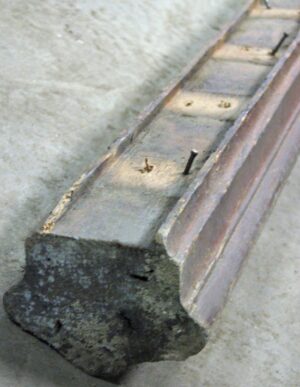 Close-up of a reclaimed wooden banister handrail with a dark wood tone, showing the carved end and visible nail holes on the top surface.