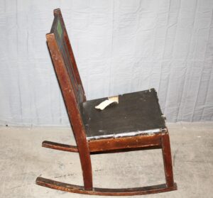 Side view of an Arts and Crafts wooden rocking chair with a black seat and dark wood frame, showing the curved rockers and simple armrests in a restorable condition.