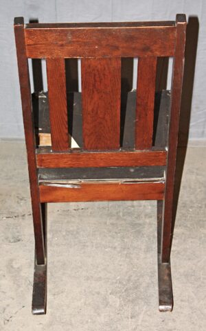 Rear view of an Arts and Crafts wooden rocker with dark wood tone, showing the slatted back and tapered legs in a simple, sturdy design.