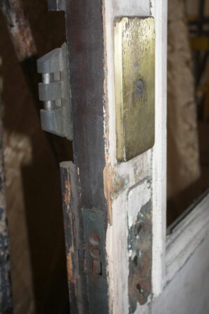 Close-up of a white entry door edge showing the hinge hardware, lock plate, and weathered wood around the missing glass opening.