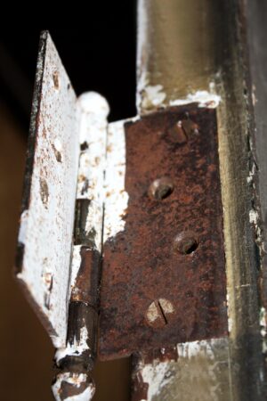 Close-up of a vintage French passage door hinge and latch area, showing peeling paint, rust, and the edge of one door panel in dark wood tone.