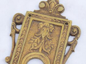 Close-up of a French-style bronze door back plate with a keyhole opening, ornate scroll edges, and a decorative floral relief.