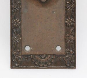 Neoclassical bronze door pull plate with floral and shell border detail, two mounting holes, and a visible bent center plate.
