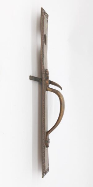 Side view of an antique bronze entry door pull with a floral plate, curved handle, thumb latch, and lock insert, shown with the plate bent.