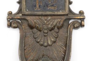 Close-up of an ornate bronze door knocker with a removable room number plate and decorative raised scrollwork.