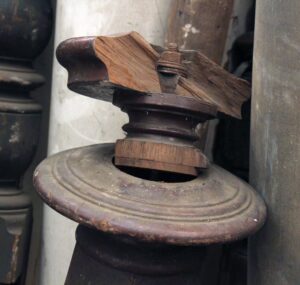 Close-up of an antique traditional newel post in a dark wood tone with a carved top piece and threaded hardware visible.