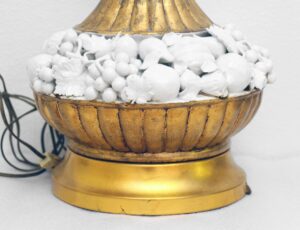 Close-up of a vintage Italian table lamp with a gilt gold base and white ceramic fruit decorations around the center, showing the lower body and decorative details.