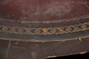 Close-up of a round wooden side table with a worn leather inlaid top and decorative trim, showing edge chipping and surface scuffs.