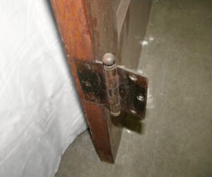 Close-up of a pine closet door hinge on a narrow interior door, showing the dark wood tone side and a crackled painted edge.