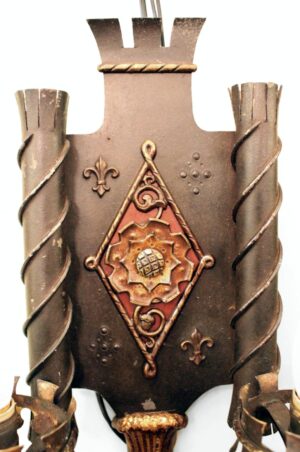 Close-up of a Tudor-style wall sconce with ornate bronze details and wrought iron side scrolls, shown from the front.