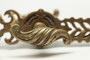 Close-up of an antique bronze door handle with a serpentine lever, showing decorative cutout back plate details in the background.