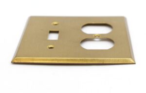 Brushed brass wall plate for a toggle switch and duplex outlet, shown at an angle with visible surface wear and cutouts for both openings.
