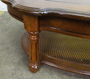 Close-up of an oval coffee table with a glass top, a turned post leg, and a lower shelf with a caning pattern.