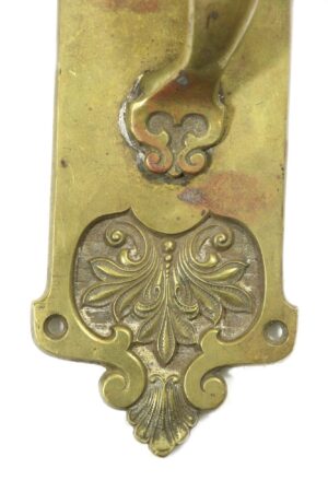 Close-up of a Romanesque brass door pull showing the decorative lower plate and thumb insert area, with the lock hole visible and the thumb latch missing.
