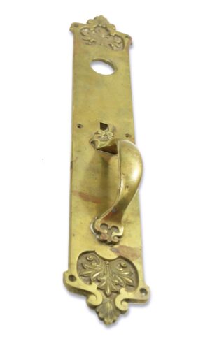 Romanesque brass door pull with an oval lock hole and ornate thumb insert, shown vertically with the thumb latch missing.