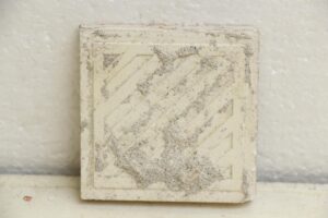 Square ceramic wall tile with a textured surface and age-related chips and scrapes, shown in close-up.