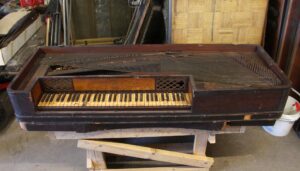 Antique piano forte keyboard in dark wood tone, shown with the exposed keys and strings of the instrument’s upper section.