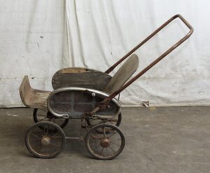 Vintage baby stroller with a medium wood tone body and four small wheels, shown in profile with a long curved metal handle and Victorian-style construction.