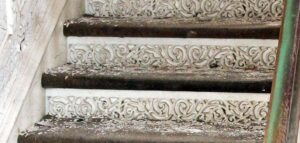 Close-up of a white cast iron staircase with ornate scrollwork risers and dark worn treads, shown from the left side with the railing on the right.