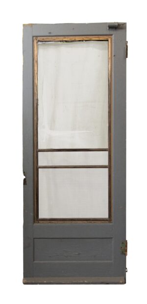 Old screen door with a large upper panel, lower panel, and intact hardware, shown in gray paint with a screened center section and worn wood trim visible.